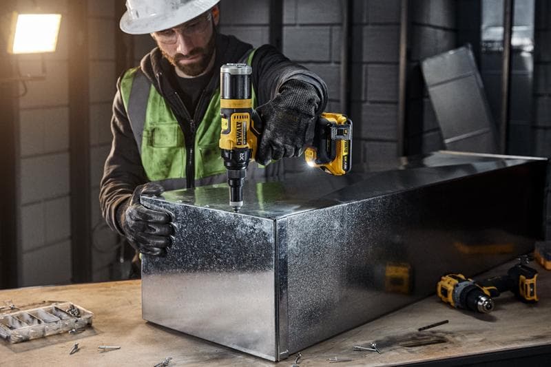 O persoană echipată cu echipament de protecție folosește uneltele electrice fără fir DEWALT DCF403NT pentru a lucra la o cutie metalică într-un atelier industrial. Diverse șuruburi și unelte sunt vizibile pe bancul de lucru.