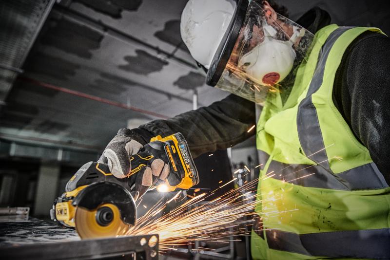 O persoană echipată cu echipament de siguranță, inclusiv cască, vizieră, mască, mănuși și vestă de mare vizibilitate, folosește unelte electrice fără fir DEWALT DCS438N pentru a tăia metal, generând scântei.